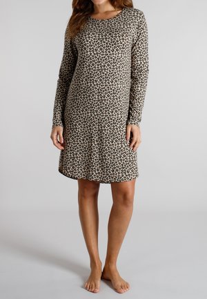 Frau trägt ein beige langärmliges Kleid mit schwarzem und braunem Leopardenmuster, steht barfuß vor einem schlichten grauen Hintergrund.