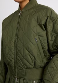 Prošívaná bomber bunda v olivově zelené barvě, se žebrovaným límcem, zapínáním na zip a bočními kapsami s logo akcentem.