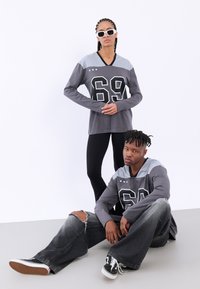 Grå langermede skjorter med "69" trykk og kontrasterende grå paneler, kombinert med svarte leggings og slitte grå jeans. Tilbehøret inkluderer solbriller.