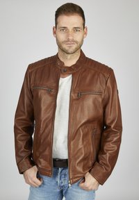 Braune Leder-Motorradjacke mit Stehkragen, gesteppten Schultern und silbernen Reißverschlüssen. Getragen über einem grauen T-Shirt und blauen Jeans.