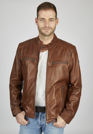 Chaqueta de motocicleta de cuero marrón con cuello levantado, hombros acolchados y cremalleras plateadas. Lleva puesta una camiseta gris y jeans azules.