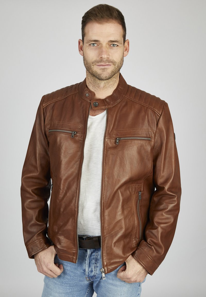 Braune Leder-Motorradjacke mit Stehkragen, gesteppten Schultern und silbernen Reißverschlüssen. Getragen über einem grauen T-Shirt und blauen Jeans.