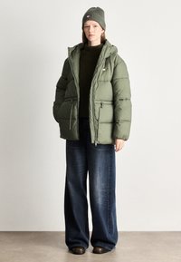 Giacca a vento verde oliva con cappuccio, tasche con zip e una texture sottile, abbinata a jeans larghi scuri e un berretto in maglia abbinato.