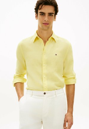 Mężczyzna o brązowych włosach, ubrany w żółtą koszulę na guziki z długim rękawem i białe spodnie, stoi z rękami w kieszeniach.