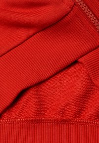 Tessuto rosso strutturato con sezioni lisce e rigate, dotato di una zip visibile lungo il bordo. Nessun motivo aggiuntivo presente.