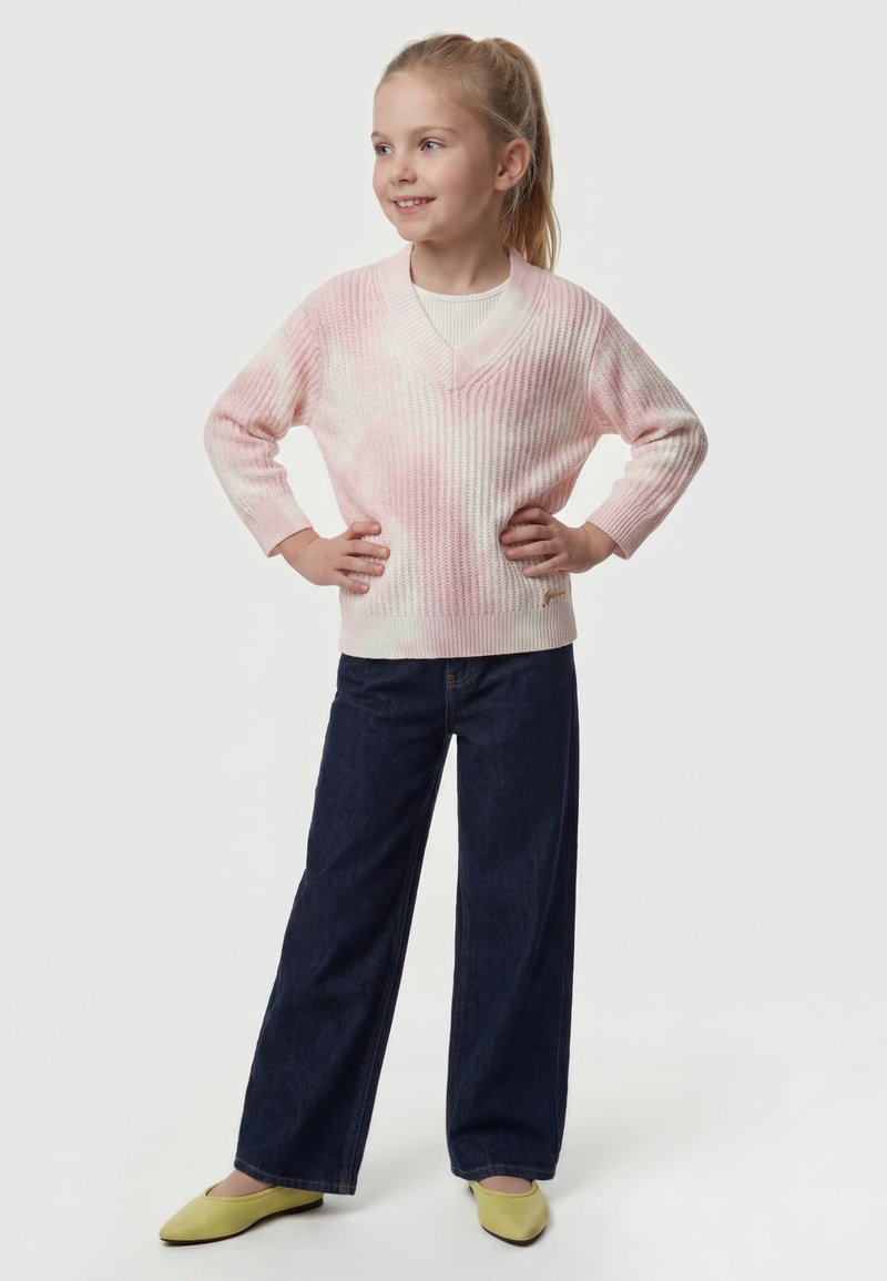 Jeune fille debout, mains sur les hanches, portant un pull rose, un jean large foncé et des chaussures vert clair sur fond blanc.