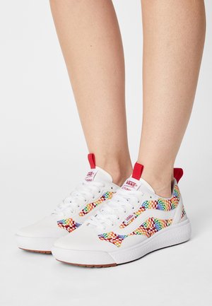 Zapatillas Vans blancas con patrón de tablero de ajedrez multicolor, lazos rojos, usadas en los pies contra un fondo neutro.