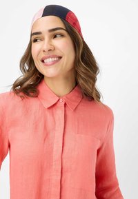 Light coral linen shirt with a collar and button placket, featuring a chest pocket. The model has a multicolored headband in shades of red and navy.
