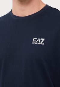 Man som bär en marinblå t-shirt med rund halsringning med vitt EA7 Emporio Armani-logotyp på bröstet.