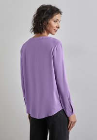 Blusa de manga larga en lavanda con escote redondo, textura suave, aberturas laterales y puños con botones. Combinada con jeans oscuros.