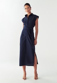 Abito camicia navy con maniche corte, bottoni sul davanti, cintura in vita, spacco laterale e texture liscia. Indossato con sandali con tacco color carne.