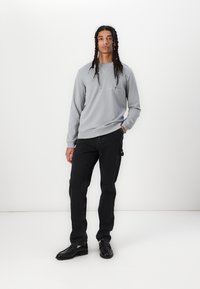 Grauer Sweater mit gerippten Bündchen und Saum, versehen mit zarter Stickerei; kombiniert mit schwarzen Jeans und schwarzen Schuhen. Das Model steht vor einem weißen Hintergrund.