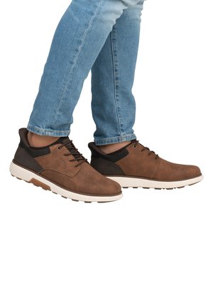 Chaussures en cuir marron avec des accents noirs, dotées d'une semelle en caoutchouc et d'un design à lacets. Portées avec un jean bleu clair, présentant des surfaces texturées.