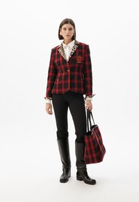 Una donna posa indossando un blazer a quadri rossi con colletto in stampa leopardata, pantaloni neri e stivali al ginocchio, mentre tiene una borsa a tracolla coordinata in plaid.