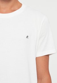 Tricou alb din bumbac cu guler rotund și mâneci scurte. Prezintă un mic "R" brodat în negru pe piept. Textură netedă.