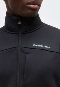 Άνδρας με μαύρο μπουφάν με φερμουάρ και ψηλό γιακά και λευκό λογότυπο "Peak Performance" στο στήθος, σε απλό φόντο.