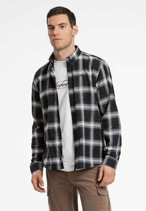 Zwart-wit geblokt flanellen shirt met knoopsluiting en verstelbare manchetten, gedragen over een witte t-shirt. Casual broeken zichtbaar daaronder.