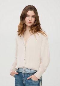 Camicia a maniche lunghe beige chiaro con scollo a V, tessuto morbido e bottoni decorativi, abbinata a jeans blu strappati.