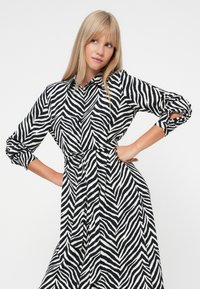 Φόρεμα με σχέδιο ζέβρας σε μαύρο και άσπρο, με γιακά, μακριά φουσκωτά μανίκια και ζώνη στη μέση, κατασκευασμένο από ελαφρύ ύφασμα.