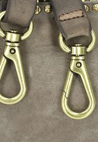 Dettaglio di una borsa in pelle grigia con grandi ganci in ottone e ganci a scatto, accentuata da borchie metalliche e cuciture marroni su una superficie liscia.