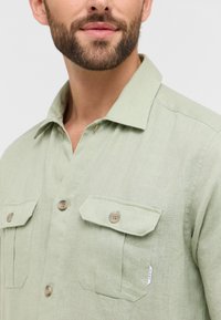 Lichtgroen overhemd van linnen, met een spread collar, twee borstzakken met klep en bruine knoopsluitingen. De textuur lijkt glad.