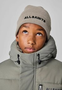 Pelēks čēpītis ar tumšu izšūtu "ALLSAINTS" tekstu, valkāts ar augstu apkakli, khaki krāsas pūkainu virsjakas, kas izceļas ar strukturētu audumu un rāvējslēdzēja akcentiem.