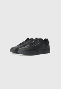 Paire de baskets basses noires Tommy Hilfiger avec lacets et étiquette de marque sur le côté, sur un fond clair uni.