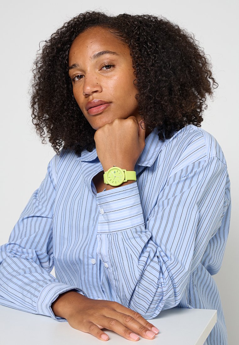Kobieta z kręconymi włosami, ubrana w niebieską w paski koszulę, opiera brodę na pięści, pokazując na swoim lewym nadgarstku limonkowy zegarek.