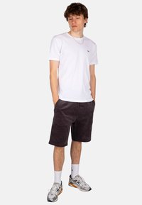 Weißes Baumwoll-T-Shirt mit kurzen Ärmeln und kleinem Logo, kombiniert mit dunkelgrauen Cordshorts. Das Modell trägt silberne Sneaker und weiße Socken.