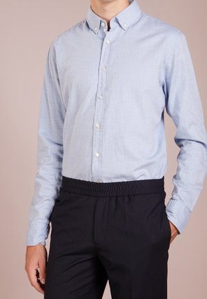 Man draagt lichtblauw overhemd met knopen, ingestopt in zwarte broek met elastische taille, staat met één hand in de zak tegen een effen achtergrond.