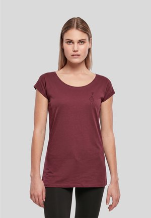 Femme aux cheveux blonds raides portant un t-shirt bordeaux à manches courtes avec un petit motif noir de main et de cœur près de l'épaule gauche.