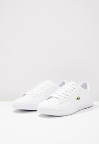 Une paire de sneakers blanches basses avec des lacets et un petit logo de crocodile vert sur le côté, posée sur une surface blanche.