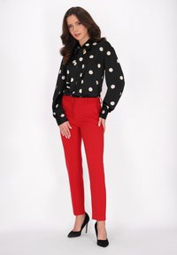 Blusa nera a pois con dettaglio del collo annodato, abbinata a pantaloni rossi aderenti e tacchi alti neri. Tessuto liscio con maniche lunghe.