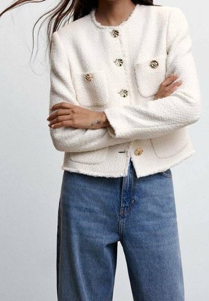 Frau trägt eine strukturierte weiße Jacke mit goldenen Knöpfen und blaue, hoch Taillierte Jeans, die Arme vor der Brust verschränkt.