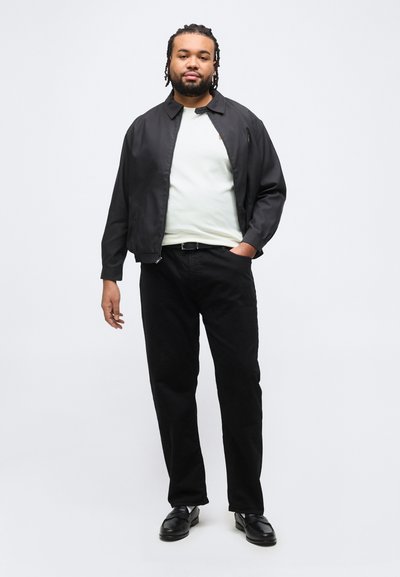 Άνδρας με κοτσίδες, που φοράει μαύρο μπουφάν, λευκό πουκάμισο, μαύρο παντελόνι και μαύρα παπούτσια, στέκεται με το ένα χέρι στην τσέπη σε λευκό φόντο.