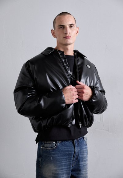 Modèle masculin portant une veste rembourrée noire brillante par-dessus une chemise noire et un jean bleu, ajustant la fermeture éclair de la veste contre un fond gris uni.