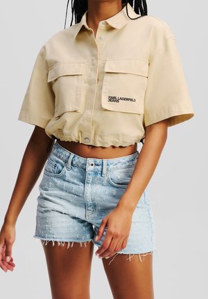Kobieta w beżowej, krótkiej koszuli z krótkim rękawem, kieszeniami z przodu i logo, w połączeniu z jasnoniebieskimi, postrzępionymi szortami jeansowymi.