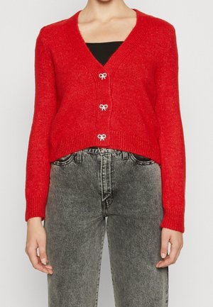 Femme portant un cardigan en maille rouge avec des boutons en forme de nœud sur un haut noir, associé à un jean gris délavé taille haute.