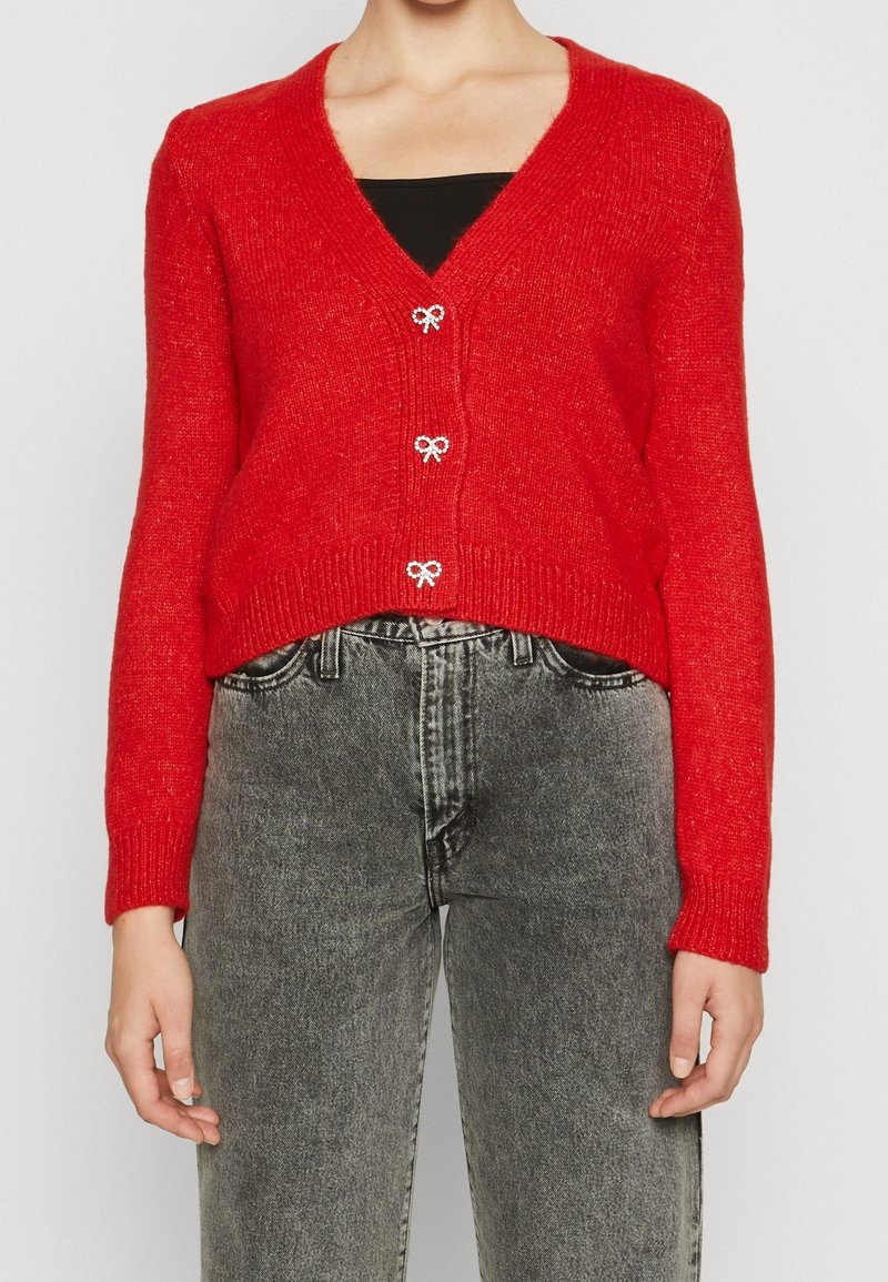 Femme portant un cardigan en maille rouge avec des boutons en forme de nœud sur un haut noir, associé à un jean gris délavé taille haute.