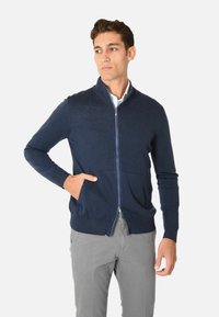 Maglione zip-up nella tonalità navy con colletto alto, polsini a costine e tasca frontale. Tessuto morbido, design aderente e materiale di peso medio.