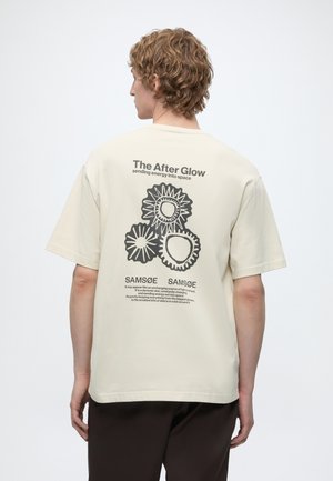 Persona con cabello rizado que lleva una camiseta beige claro con diseños negros similares al sol y texto en la parte trasera que dice "The After Glow."