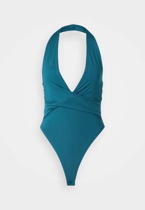 Mėlynas vientisas maudymosi kostiumėlis su gilia iškirpte, apvyniojimo dizainu ir aukštai iškirptomis kojomis. Pagamintas iš sklandžios, tamprios medžiagos. Nėra ryškių raštų ar priedų.