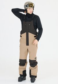 Salopette da sci con una combinazione di colori marrone chiaro e nero, dotata di spallacci regolabili, tasche con zip e materiale impermeabile per uso all'aperto.