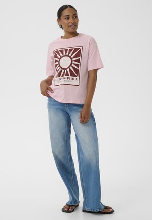 Žena v světle růžovém tričku s motivem slunce a nápisem, široké modré džíny a černé žabky stojící před jednobarevným bílým pozadím.