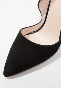 Chaussure à talon haut pointue en daim noir avec doublure intérieure beige, présentée sur un fond blanc.