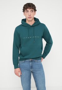 Φούτερ με κουκούλα σε τιρκουάζ χρώμα, με κορδόνια, μπροστινή τσέπη και λευκό κείμενο "JACK & JONES". Συνδυασμένο με μπλε τζιν, το μοντέλο στέκεται μπροστά σε απλό λευκό φόντο.