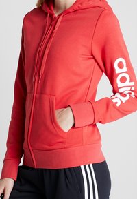 Pull rouge avec une poche avant, capuche à cordon et logo blanc sur la manche. Fabriqué en tissu doux avec un design ajusté.