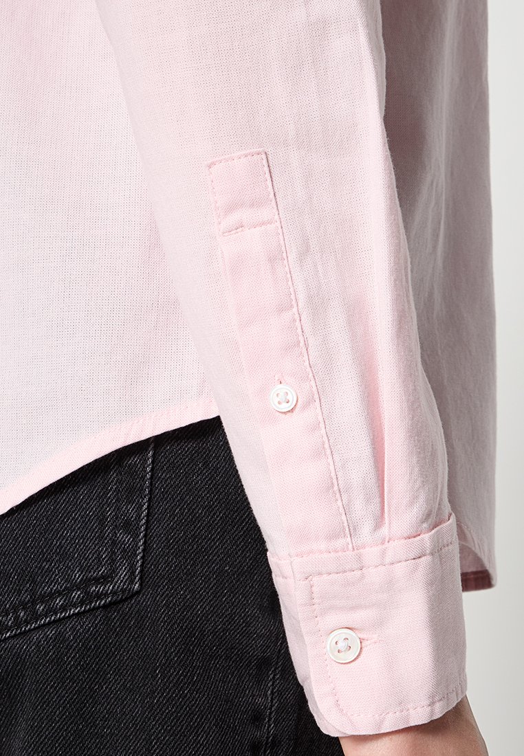 Camisa de manga larga rosa claro con tejido texturizado, puños abotonados y dobladillo curvado, combinada con pantalones de mezclilla negros.