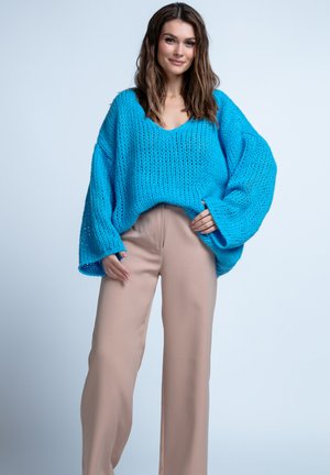 Niebieski, dziany, oversize sweter z głębokim dekoltem w kształcie litery V, teksturowanym wykończeniem i szerokimi rękawami, zestawiony z jasnobeżowymi spodniami w szerokim kroju.