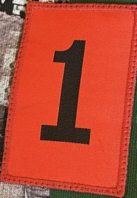 Rood stoffen label met het zwarte cijfer "1" in het midden, omrand met stiksel, gezet tegen een gestructureerde, gemusterde achtergrond.